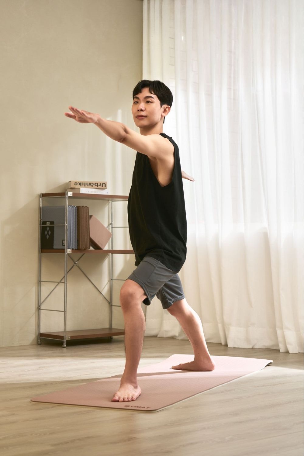Man in black tank top practicing yoga warrior pose on pink mat in bright home studio.