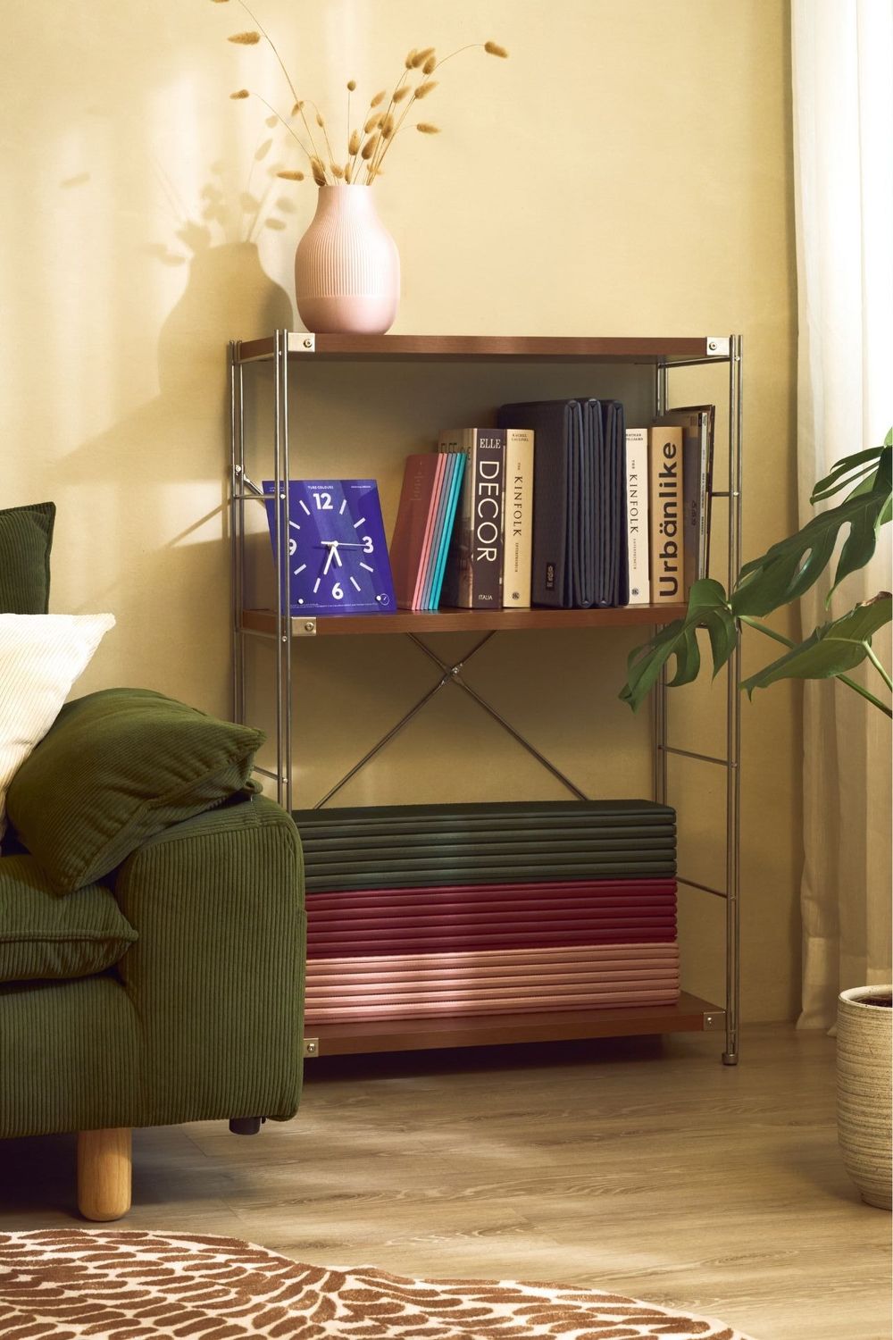 Modern bookshelf with clock, decor books, and pink vase beside green sofa in cozy living room.