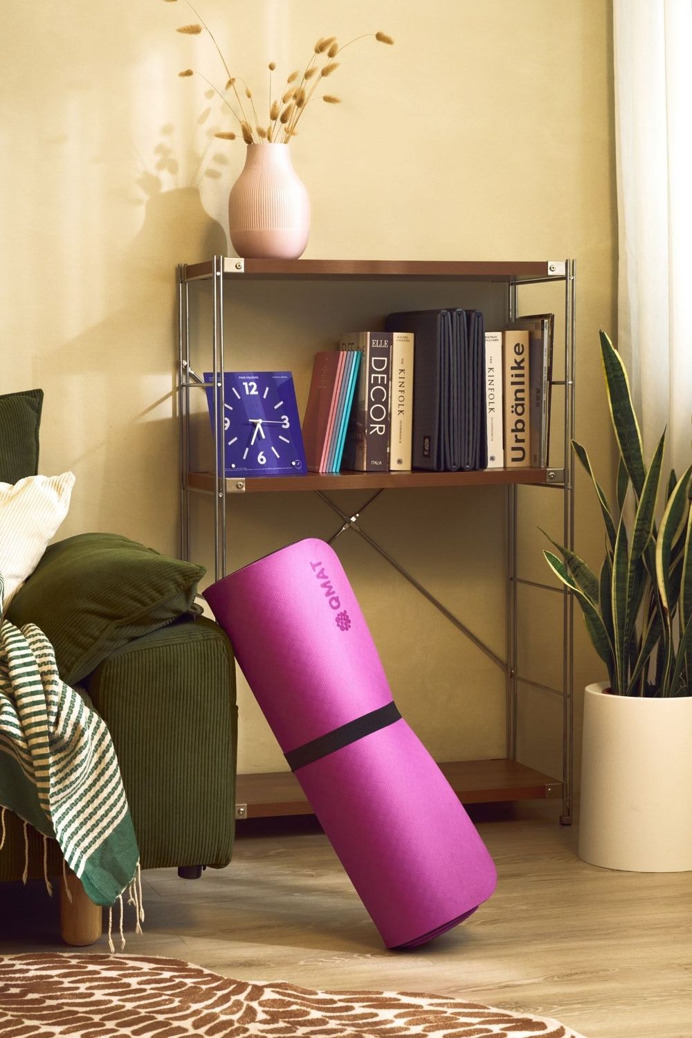 Pink rolled yoga mat leaning against a metal bookshelf in a cozy living room with books, clock, and plant decor.