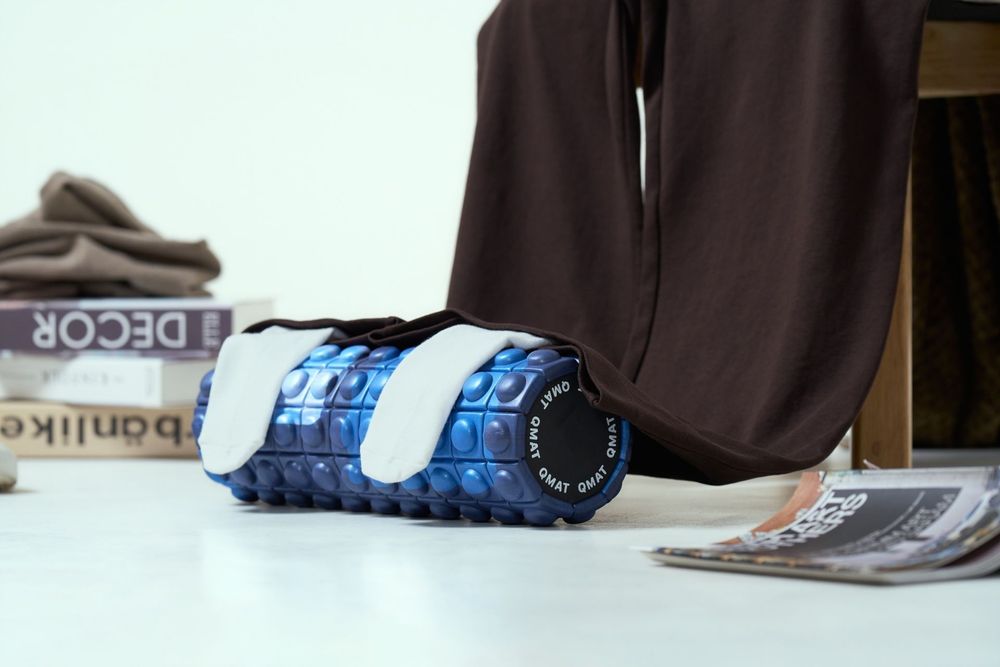 Blue textured foam roller on floor next to brown pants leg, books, and magazine in bright studio setting.