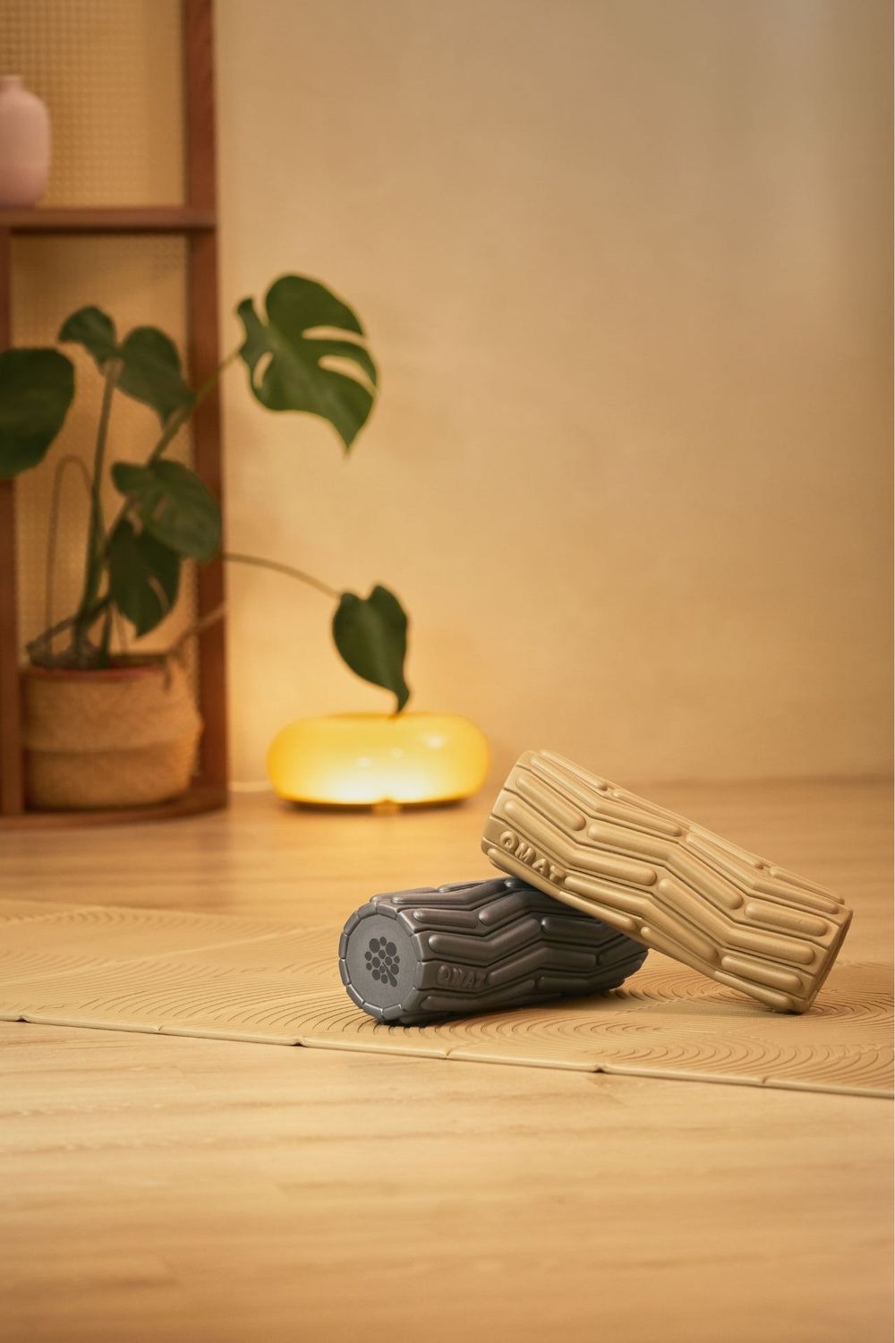 Two textured foam rollers on wooden floor with monstera plant and glowing lamp in cozy wellness room setting.