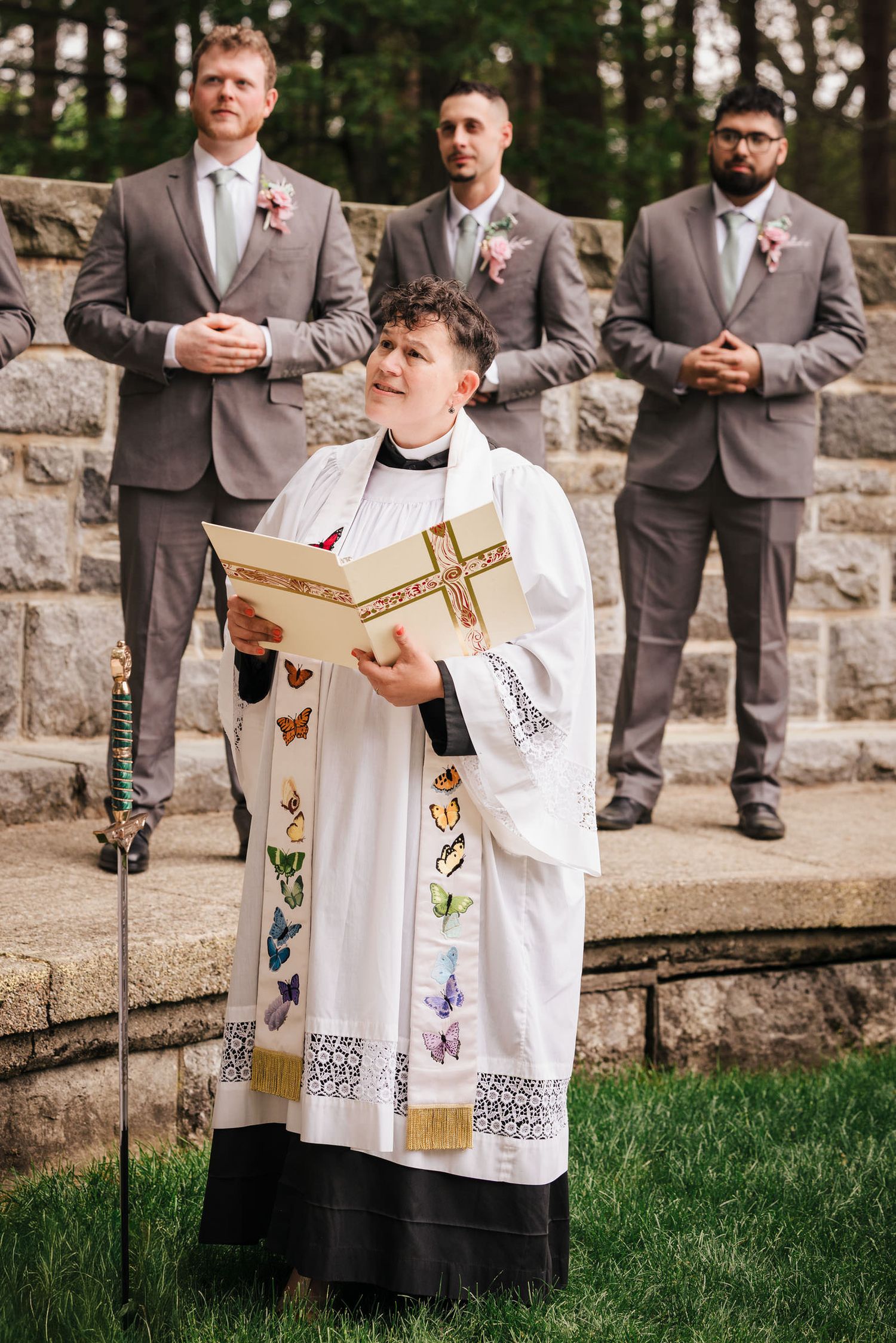 Reverend Claire reading from the ceremony book during her sermon, four groomsmen standing behind her in gray suits.