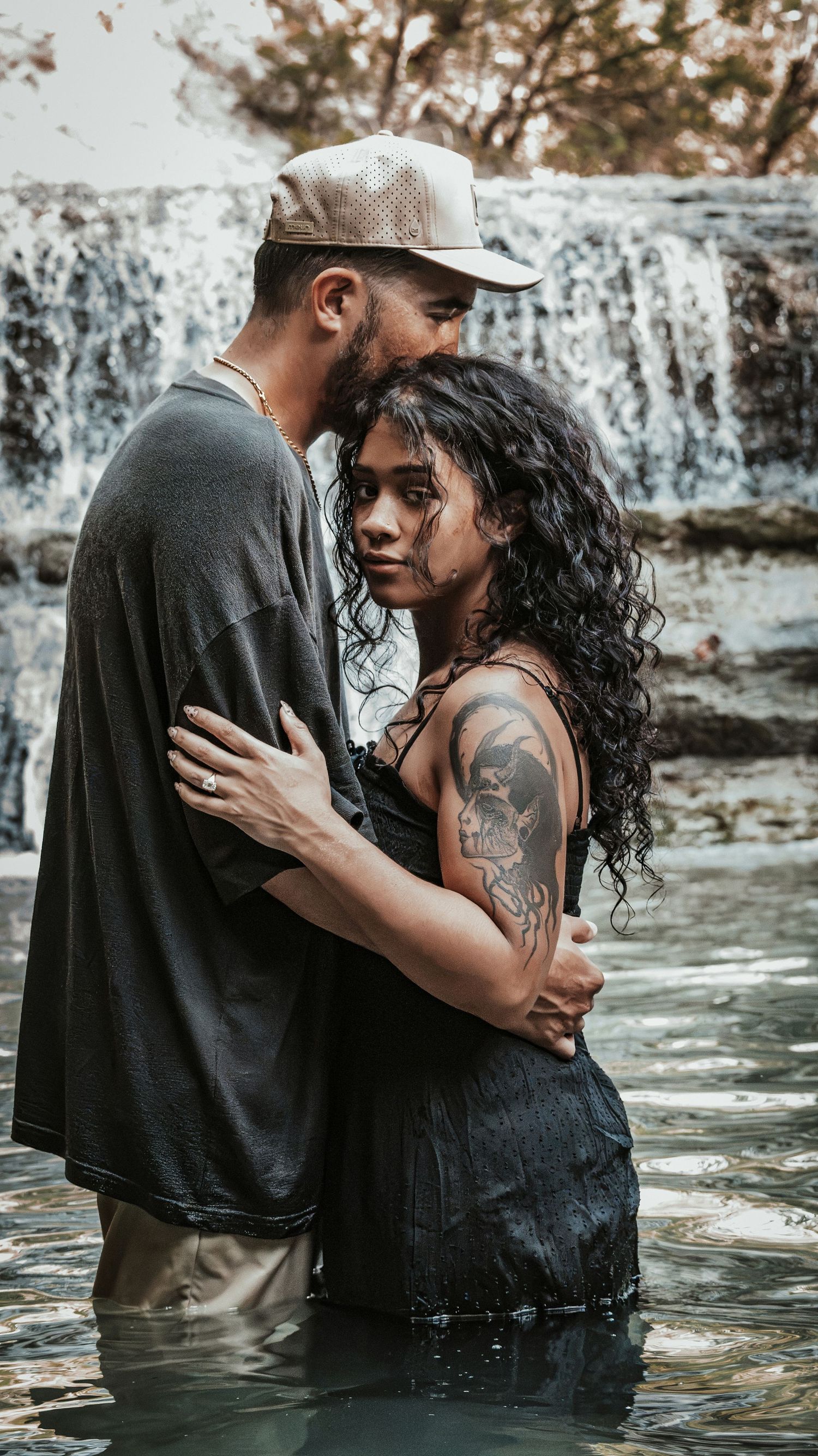 A couple embraces romantically in shallow water before a waterfall, the woman's tattoo visible on her shoulder.