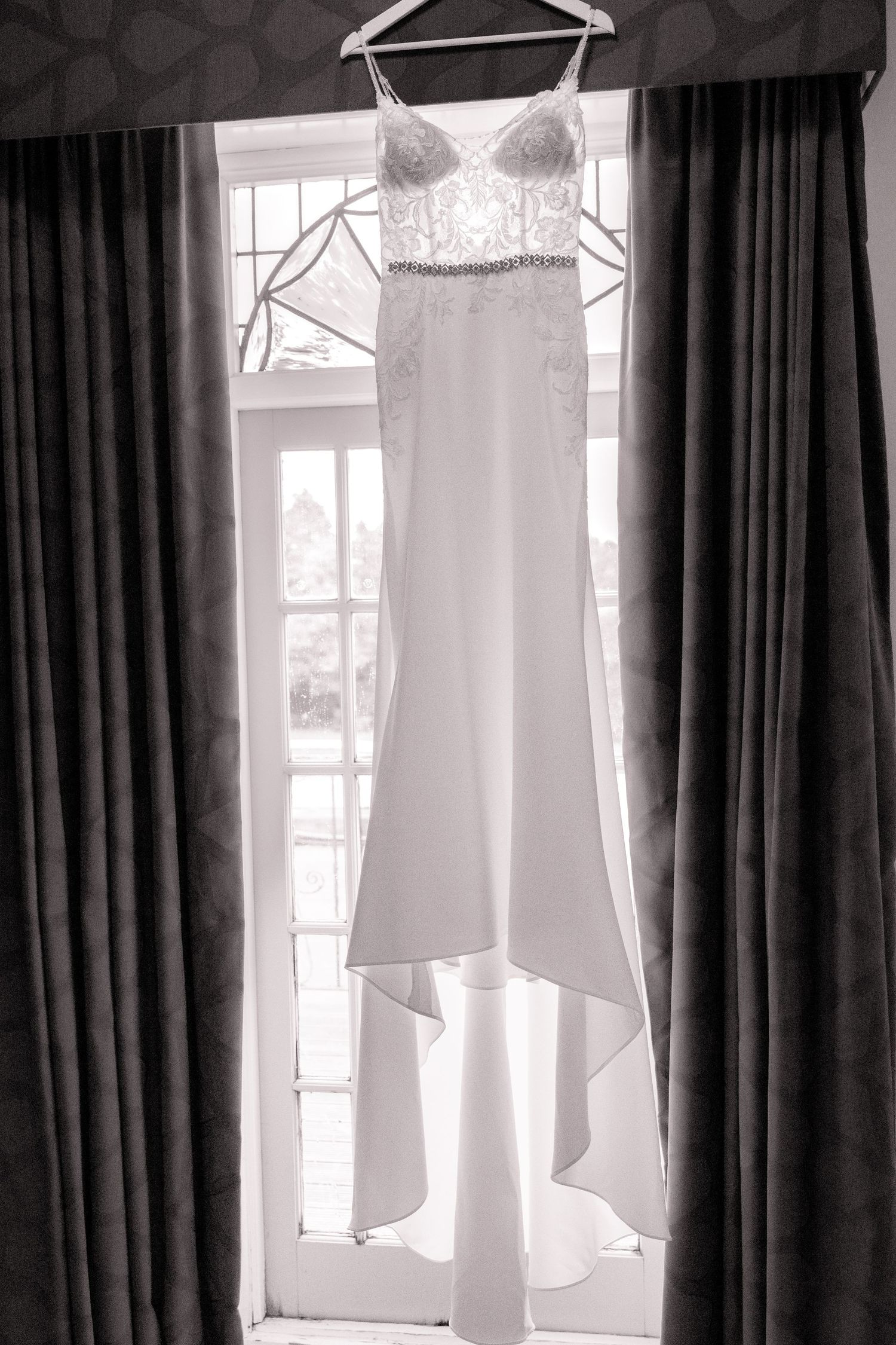 Elegant wedding dress with lace bodice hanging by a window in black and white photography.