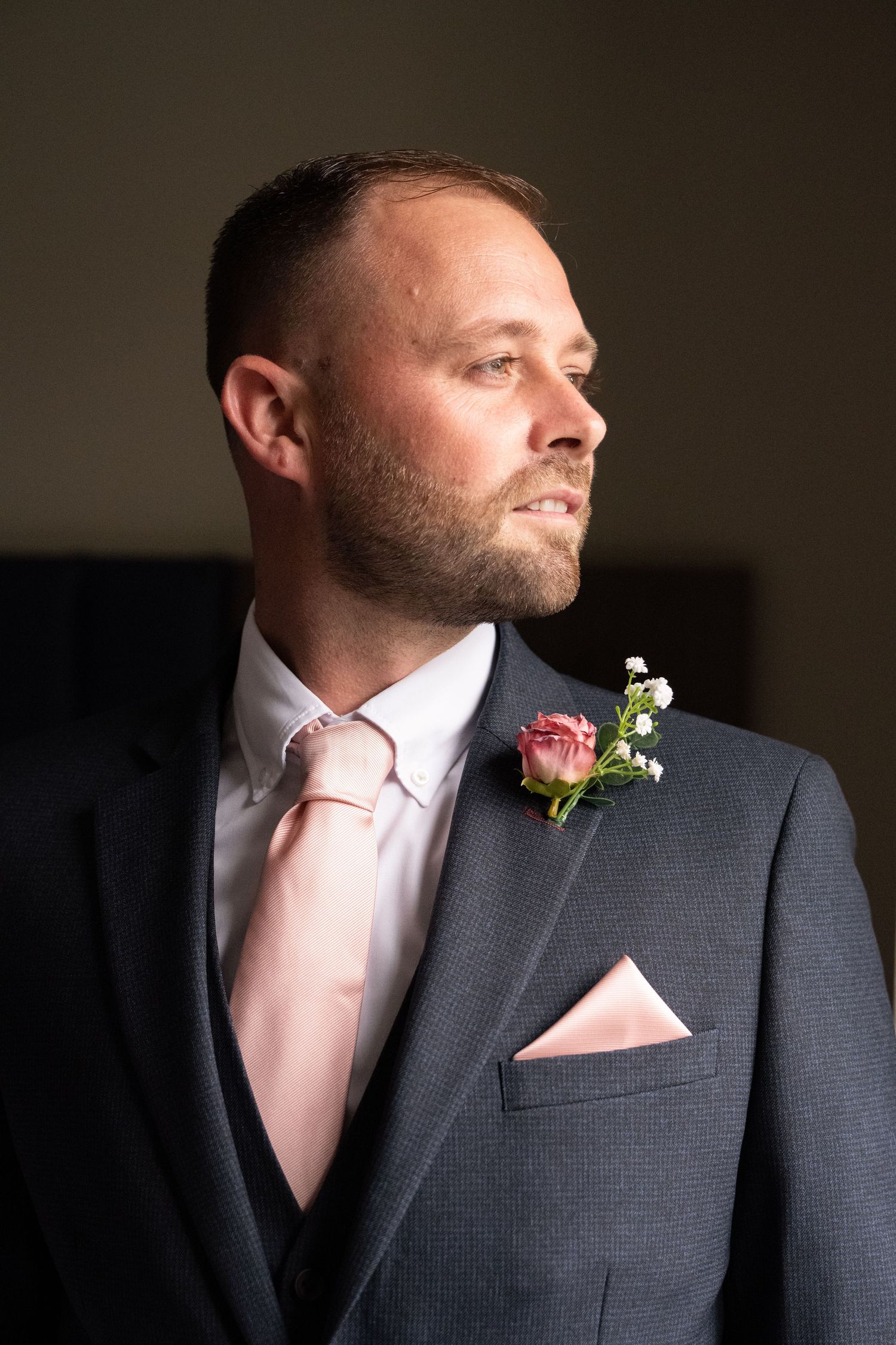 Groom in grey suit with pink tie and rose boutonniere gazing thoughtfully to the side on his wedding day.
