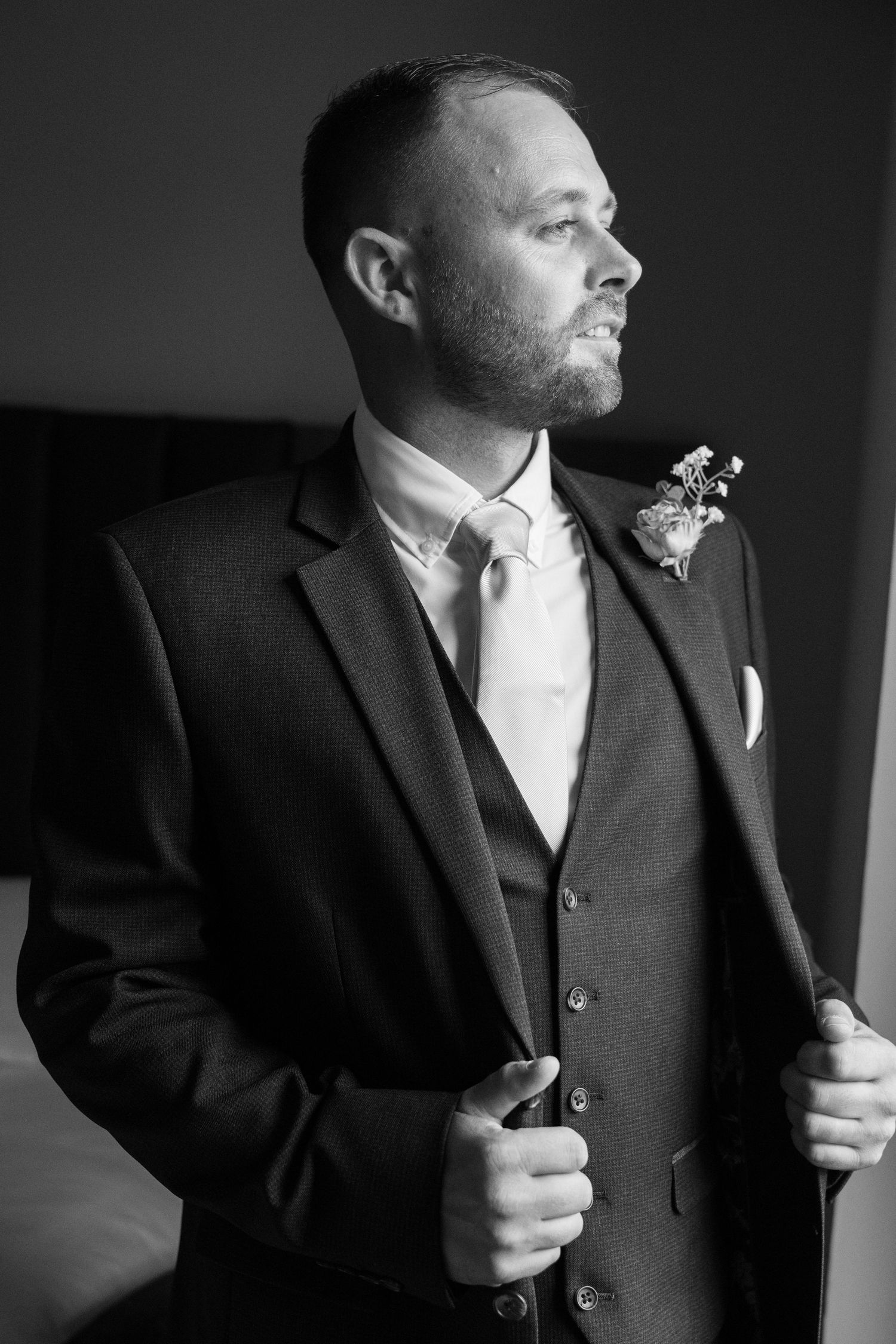 Black and white photo of a groom in a suit and boutonniere adjusting his jacket lapels before his wedding.