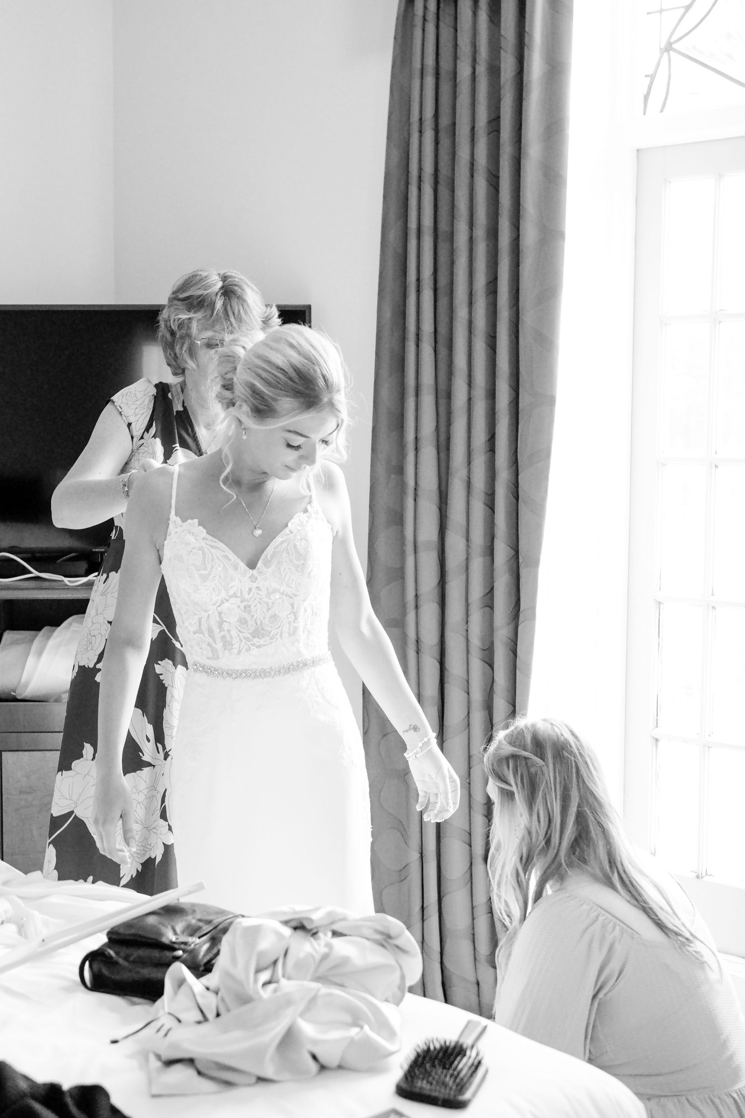 Bride being helped into her wedding dress by two bridesmaids in a bright hotel room, black and white photo.