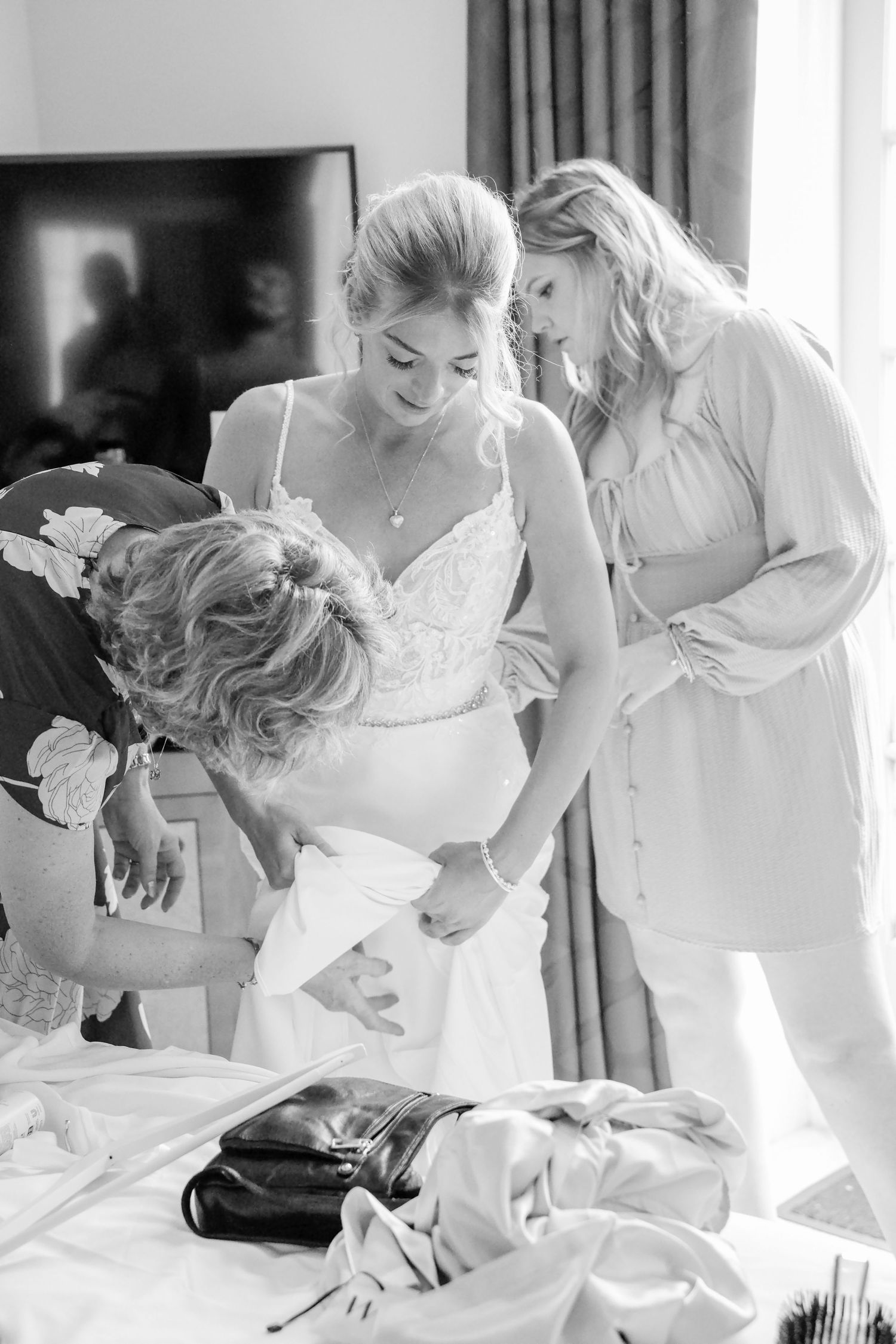Bride being helped into her white lace wedding dress by two women in a hotel room before the ceremony.