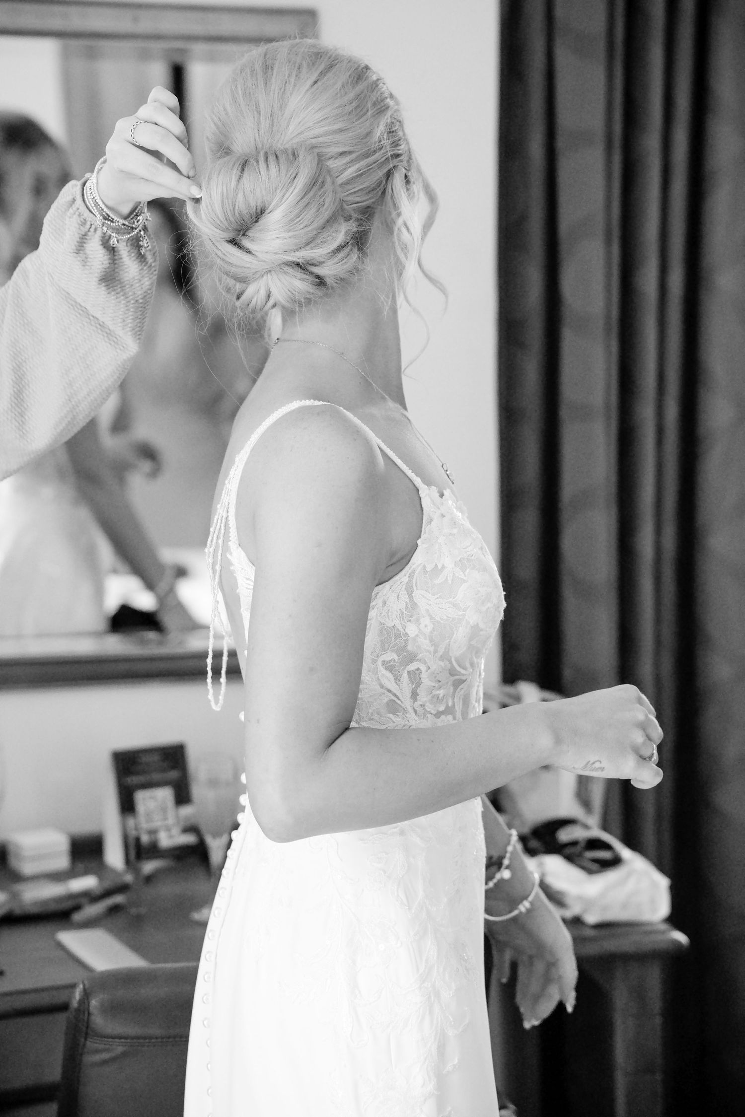 Black and white photo of bride in lace gown having hair styled before wedding ceremony.