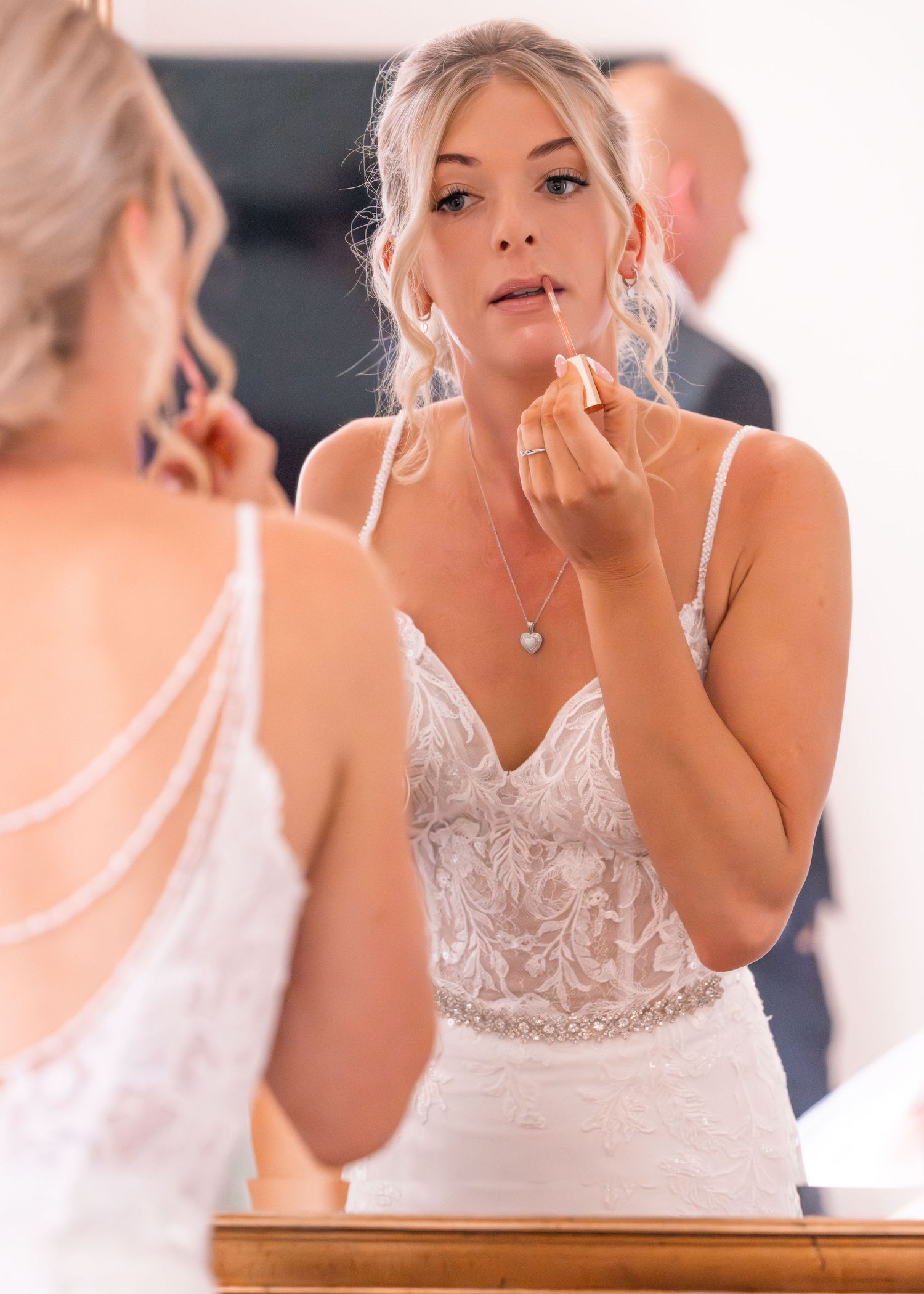 Bride in lace wedding dress applying lipstick while looking in mirror on her wedding day.