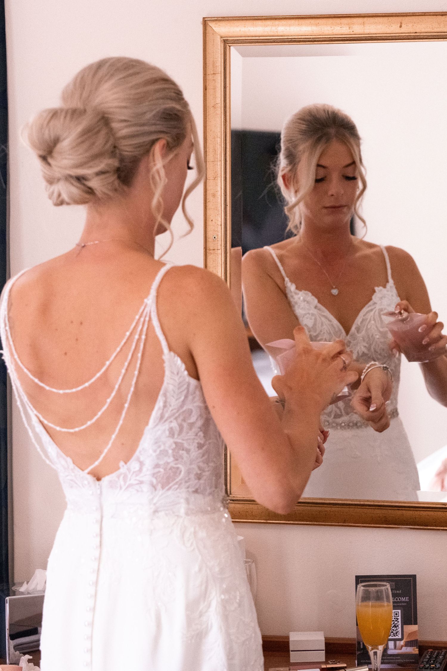 Bride in elegant backless lace wedding dress with pearl draping admires her reflection in a gold-framed mirror.