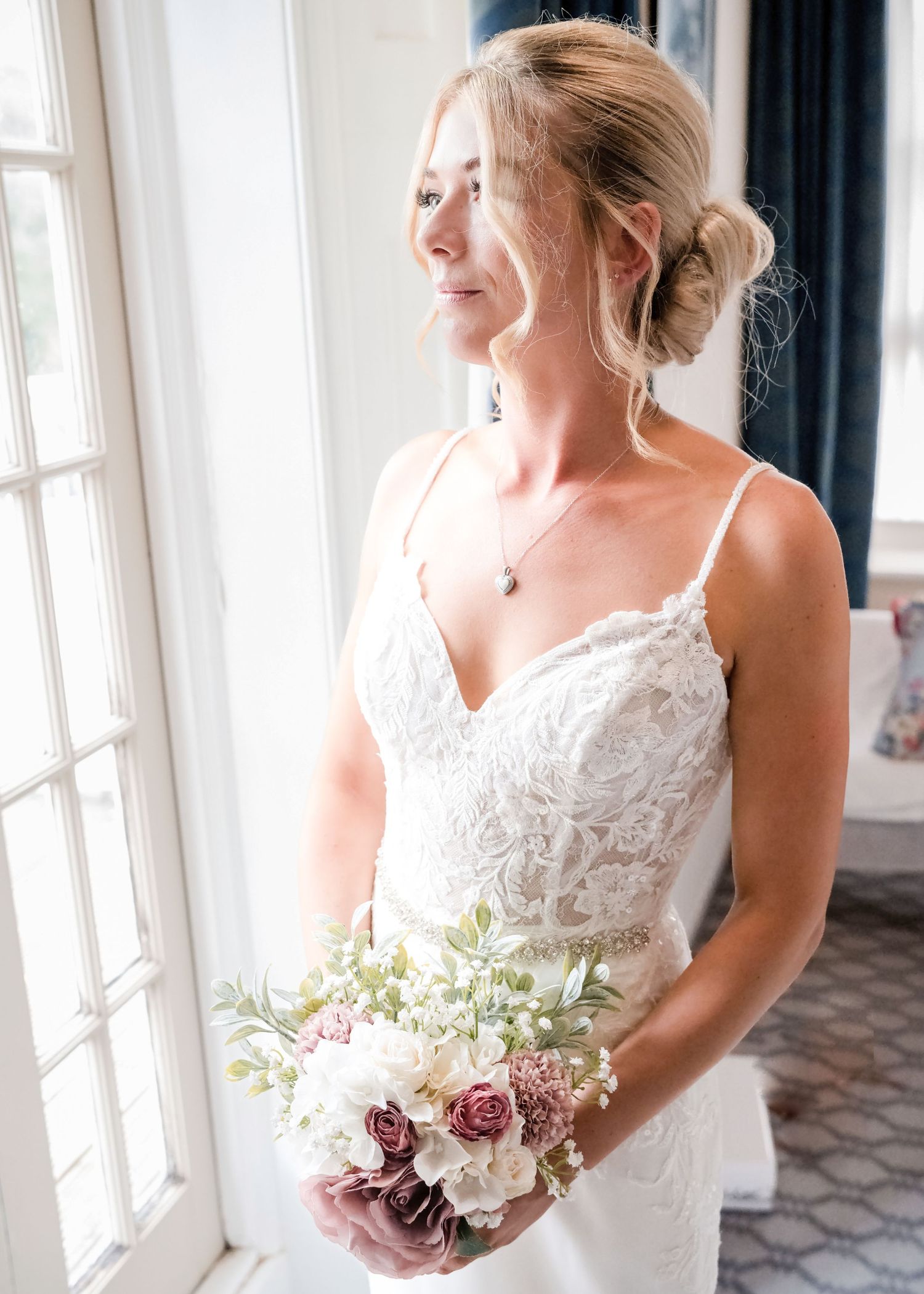 Elegant bride in lace spaghetti strap gown holding bouquet of pink roses by window.
