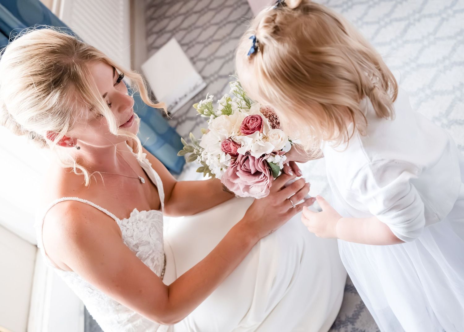 Bride in lace wedding gown smiles warmly as a young flower girl presents her with a pink bridal bouquet.