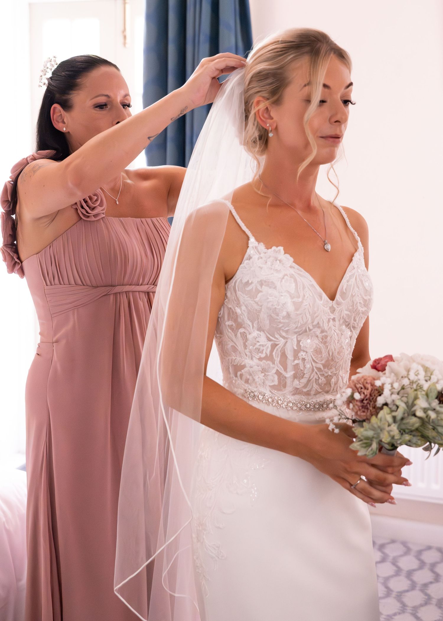Bridesmaid in pink gown adjusting veil of bride in white lace wedding dress holding bouquet.