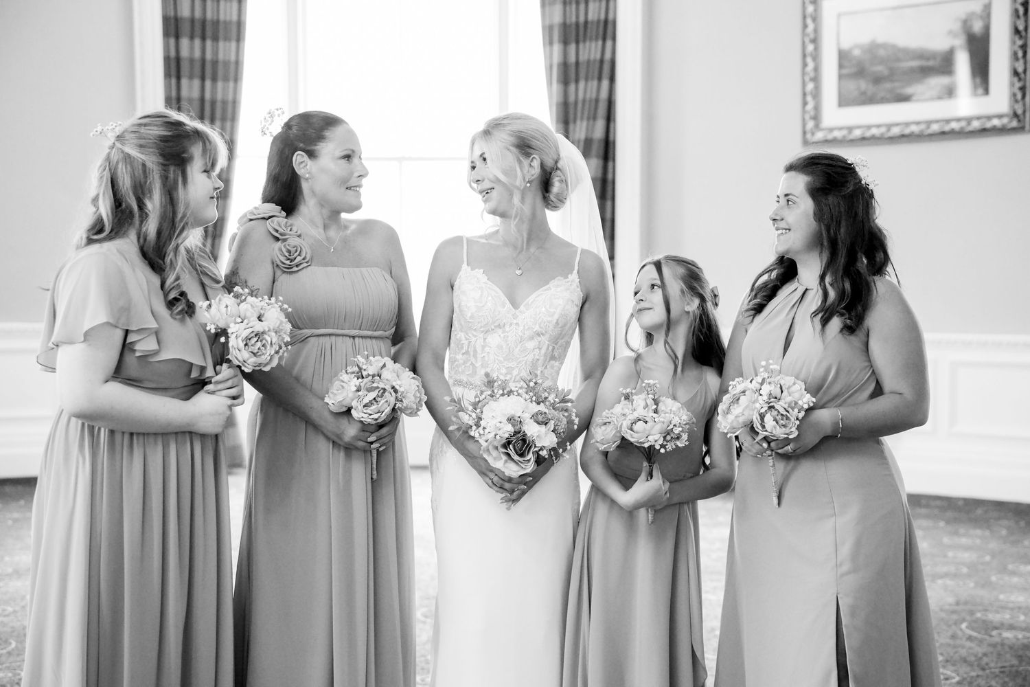 Black and white photo of a bride in a lace gown smiling with four bridesmaids in flowing dresses holding bouquets.