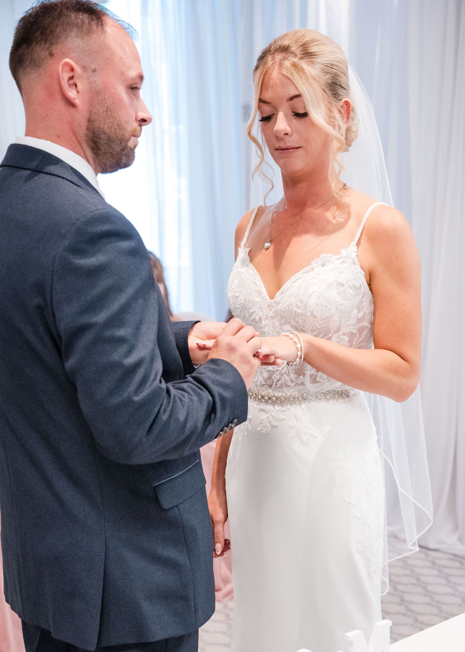 Bride placing wedding ring on grooms finger during intimate ring exchange ceremony in white venue.