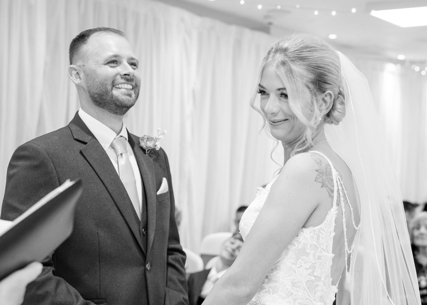 Black and white wedding photo of smiling bride and groom exchanging vows at elegant indoor ceremony.