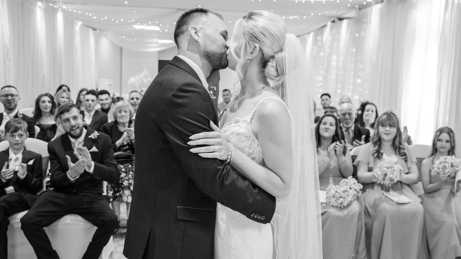 Black and white wedding photo of bride and groom sharing first kiss at altar with guests applauding.