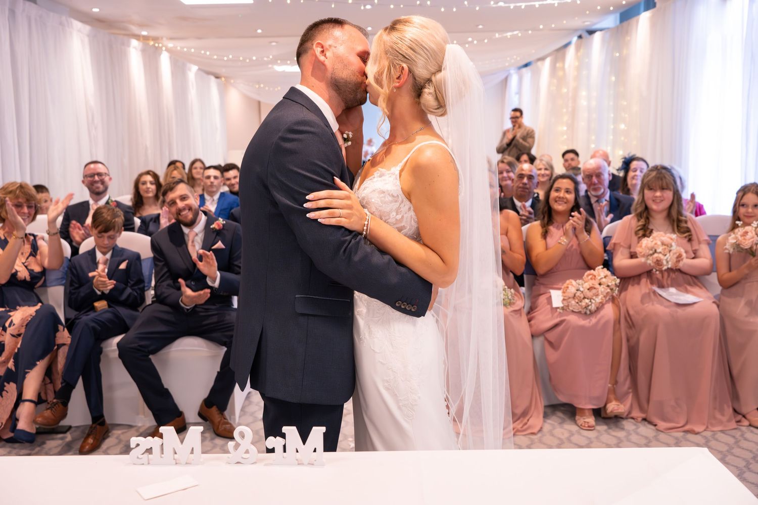 Bride and groom share first kiss at wedding ceremony with guests clapping, floral letters on table.