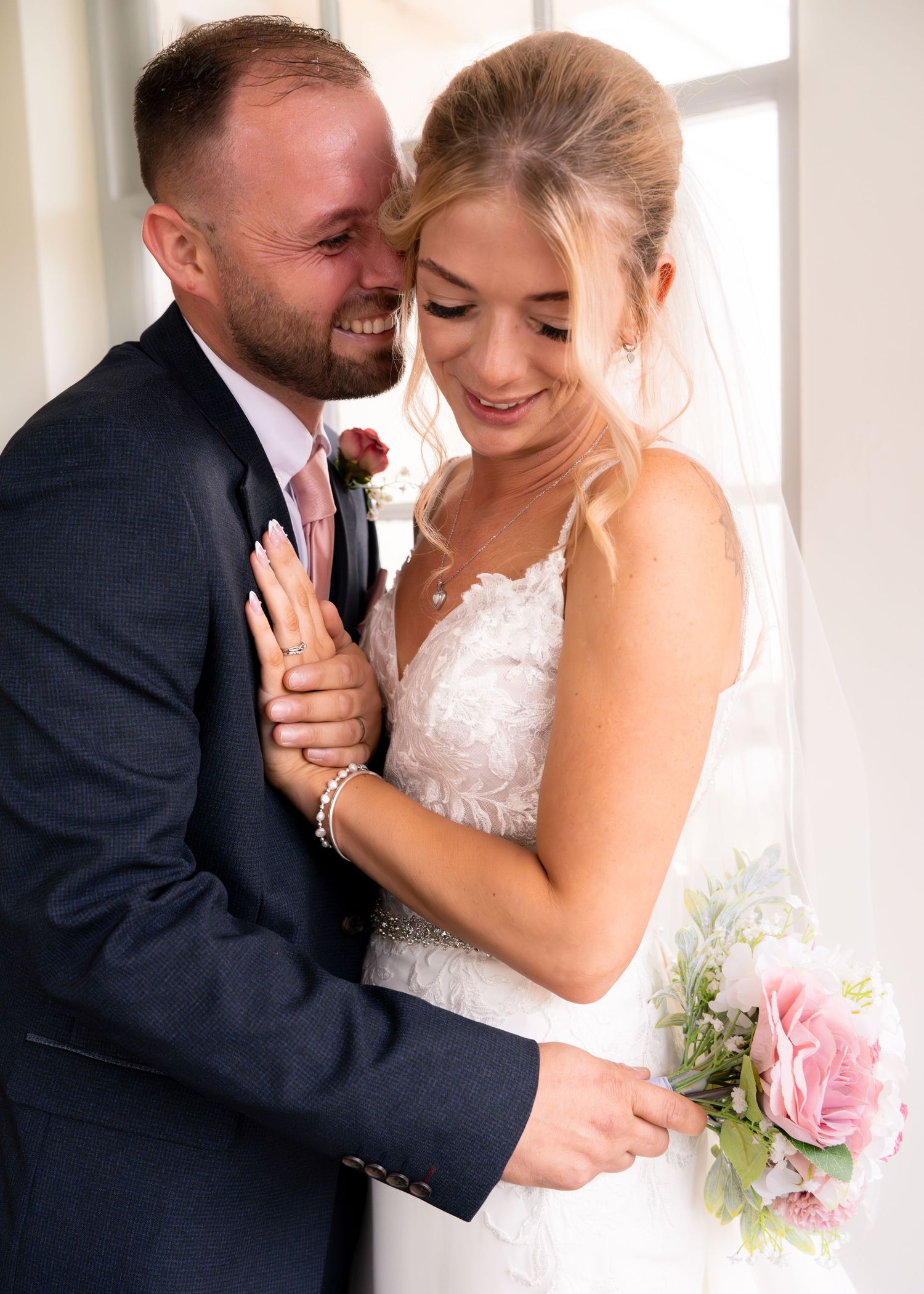 Bride and groom sharing a romantic embrace near a window, bride holding a pink rose bouquet in a lace wedding dress.