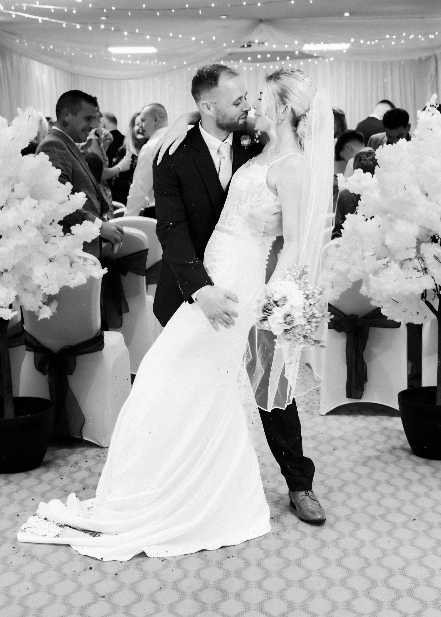 Black and white photo of newlyweds sharing first kiss as married couple in decorated wedding reception hall.