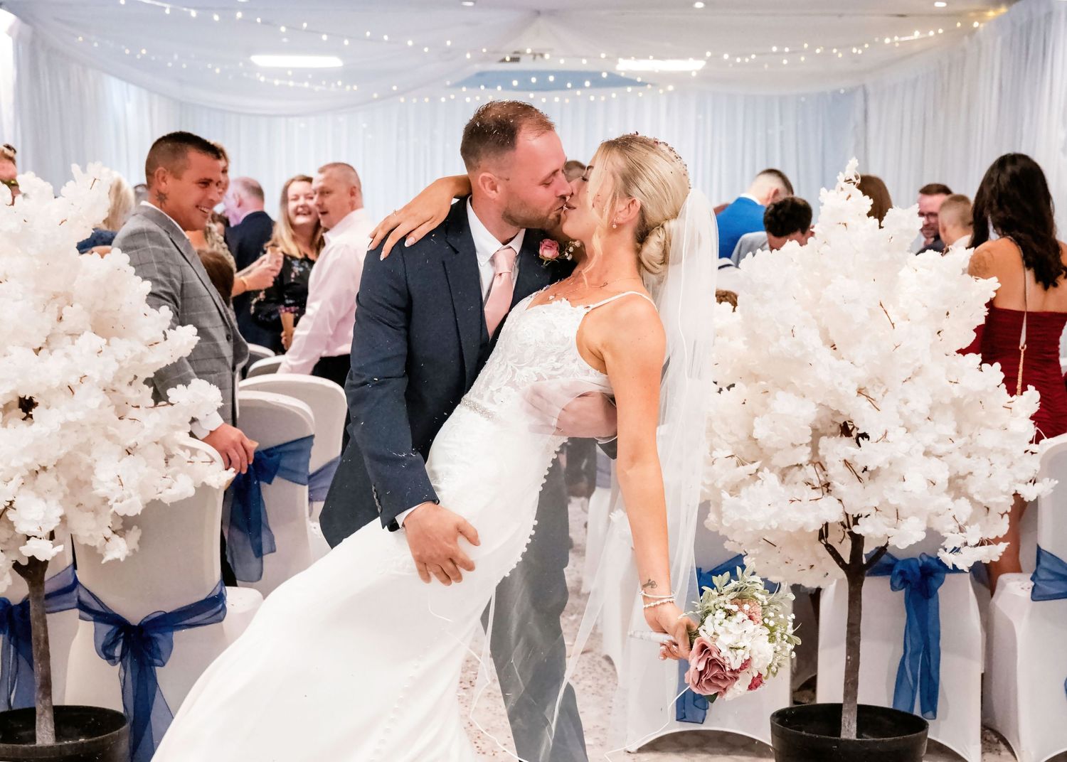 Bride and groom share a romantic kiss at their wedding reception, surrounded by white floral arrangements and blue decor.