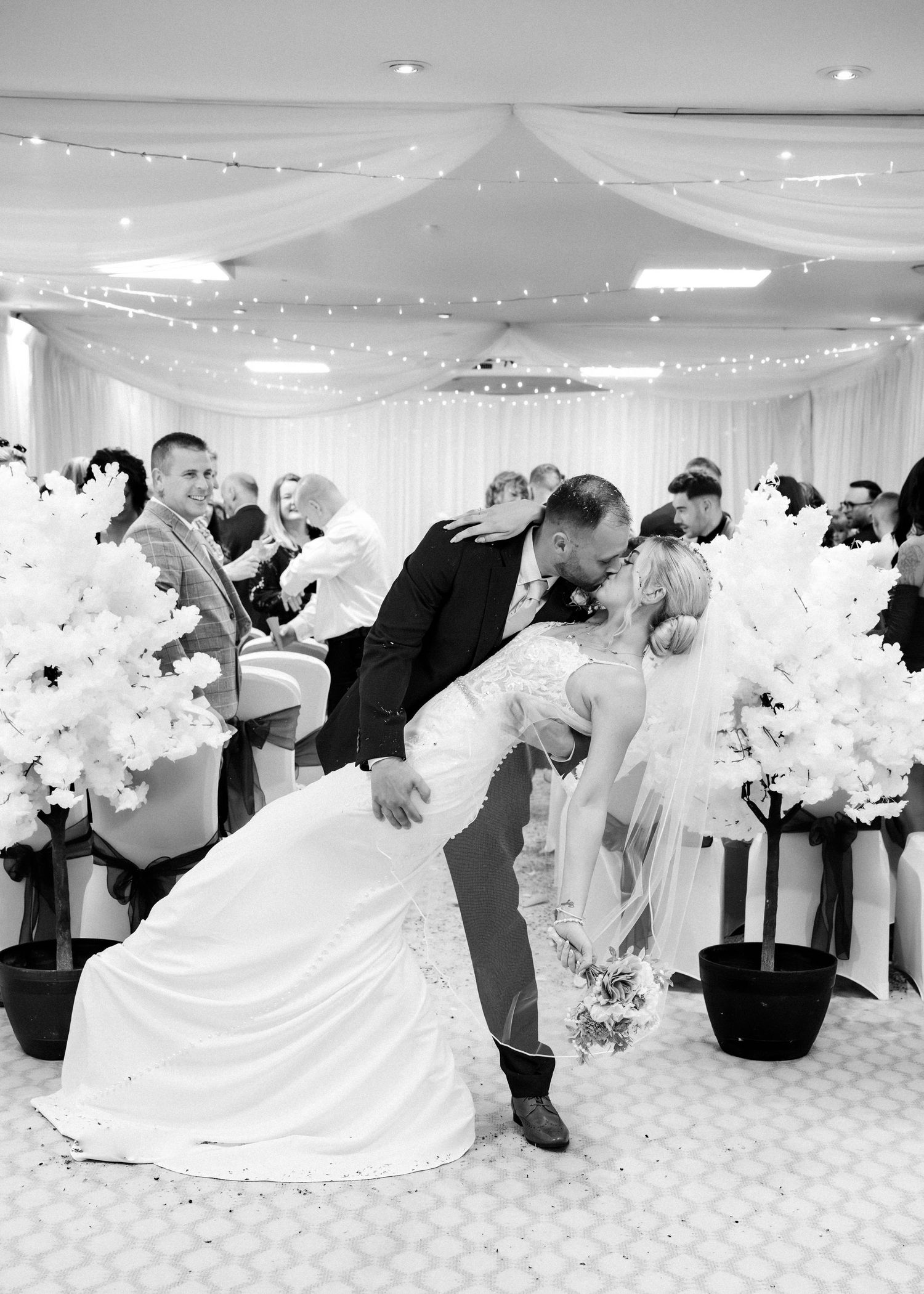 Groom dips bride for romantic kiss at wedding ceremony aisle in elegant decorated venue, black and white photo.