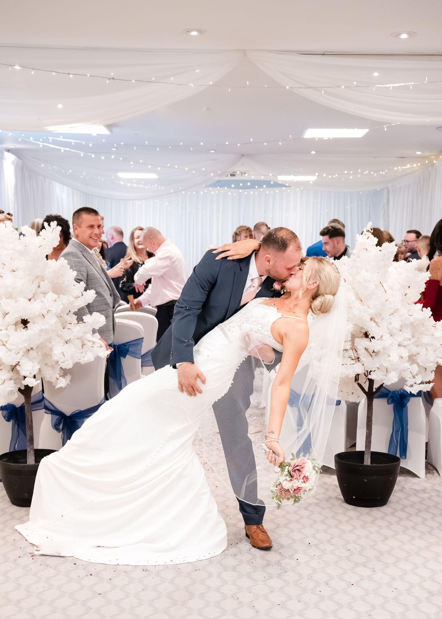 Groom dipping bride for a kiss at wedding ceremony aisle surrounded by white floral trees.