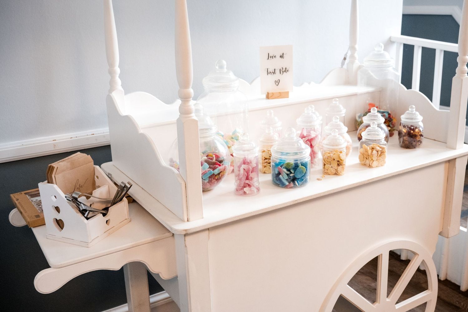 White vintage candy cart with glass jars filled with colorful sweets at a wedding reception.