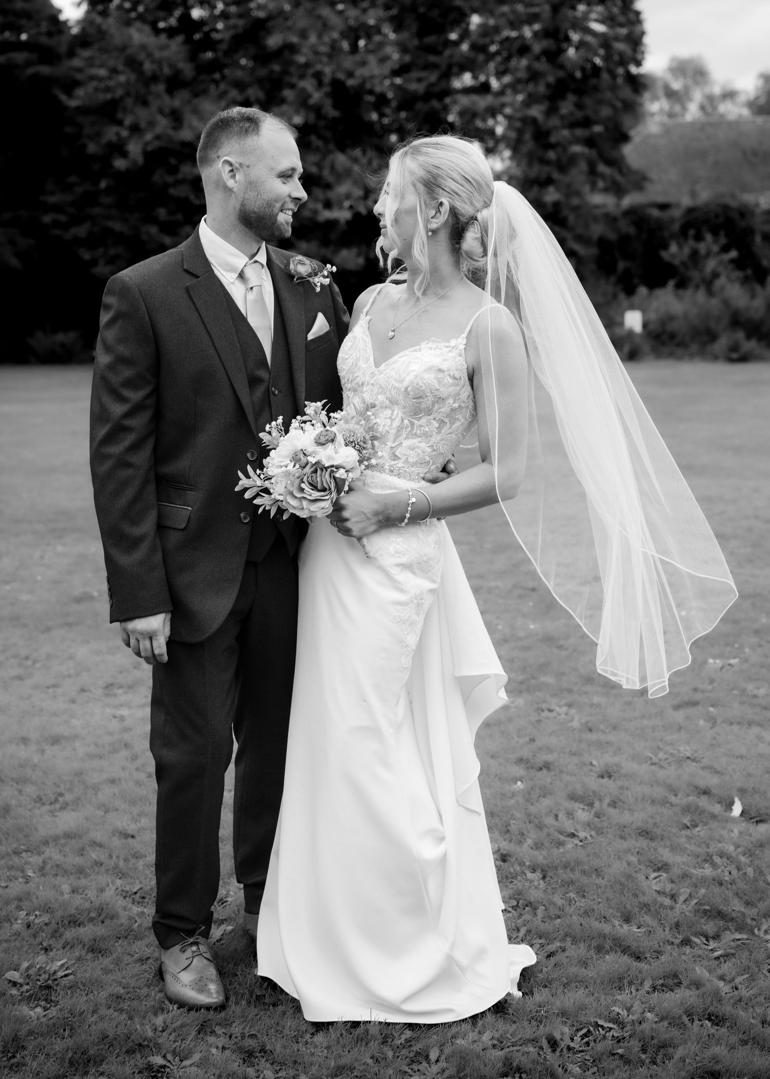 Black and white portrait of bride and groom smiling at each other in garden setting.