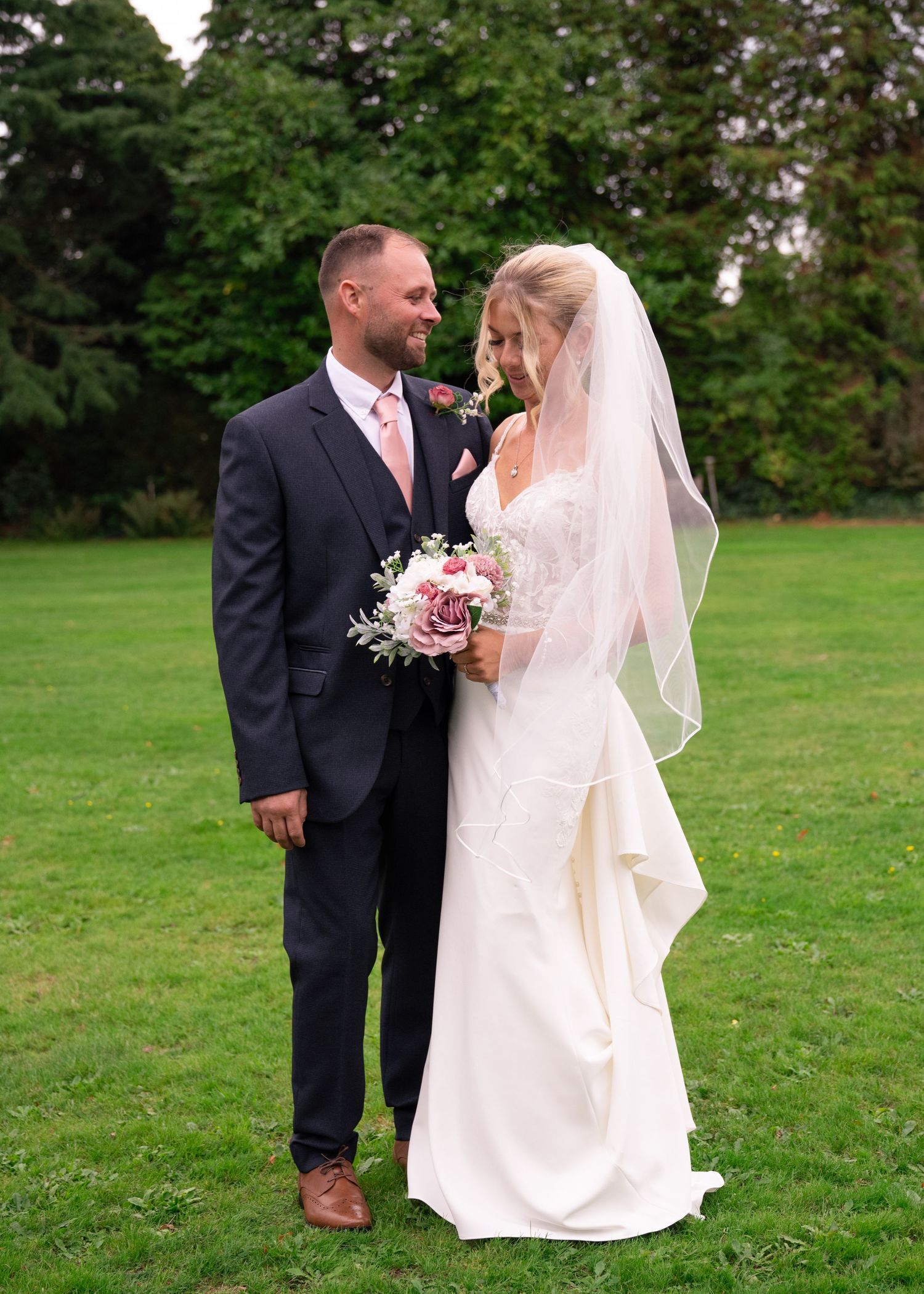 Bride and groom share a romantic moment on green lawn, bride wearing lace gown with veil holding bouquet.