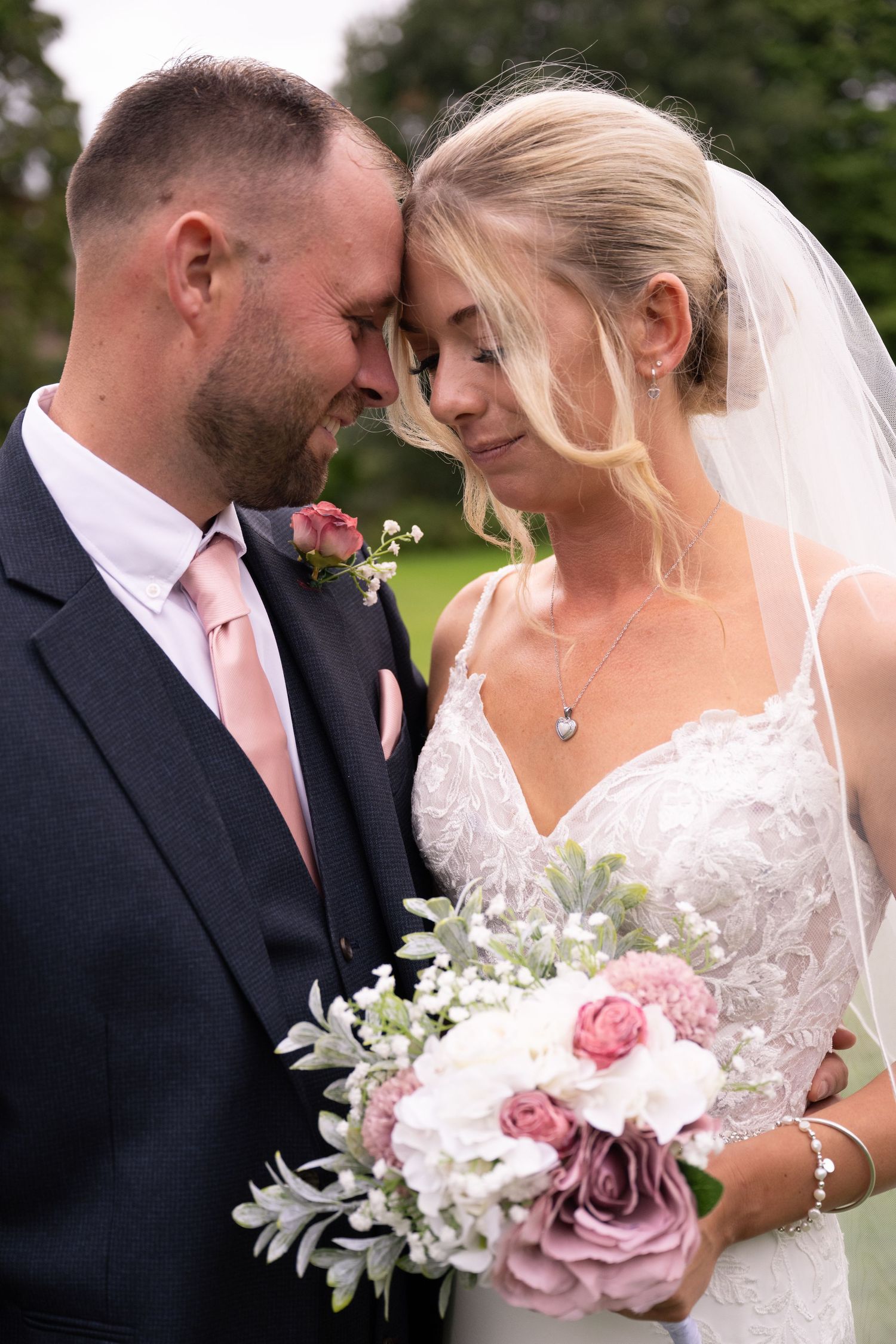 Bride and groom touching foreheads tenderly, bride holding pink and white bouquet outdoors.