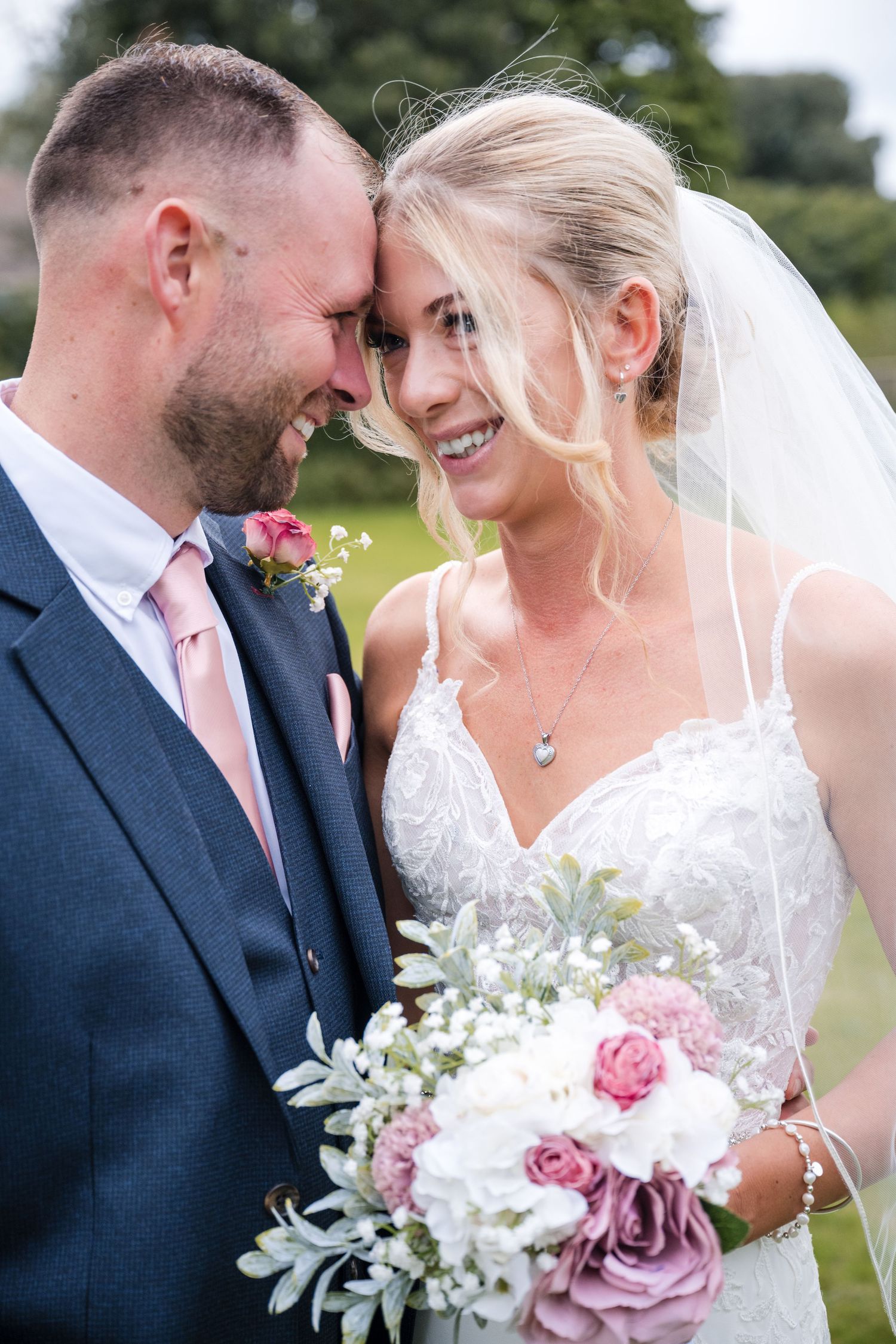 Smiling bride in lace gown with veil and groom in navy suit touching foreheads, holding pink and white wedding bouquet.