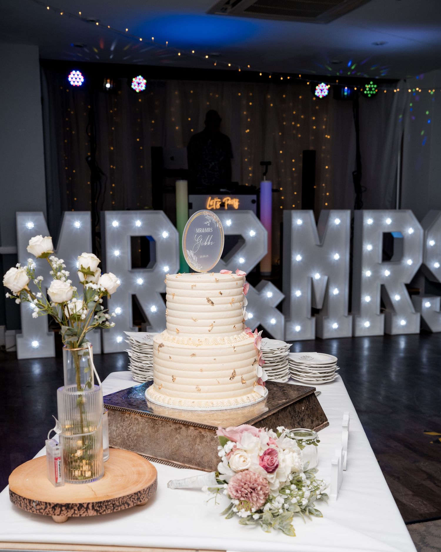 Three-tier white wedding cake displayed on wooden stand with illuminated MR & MRS letters.