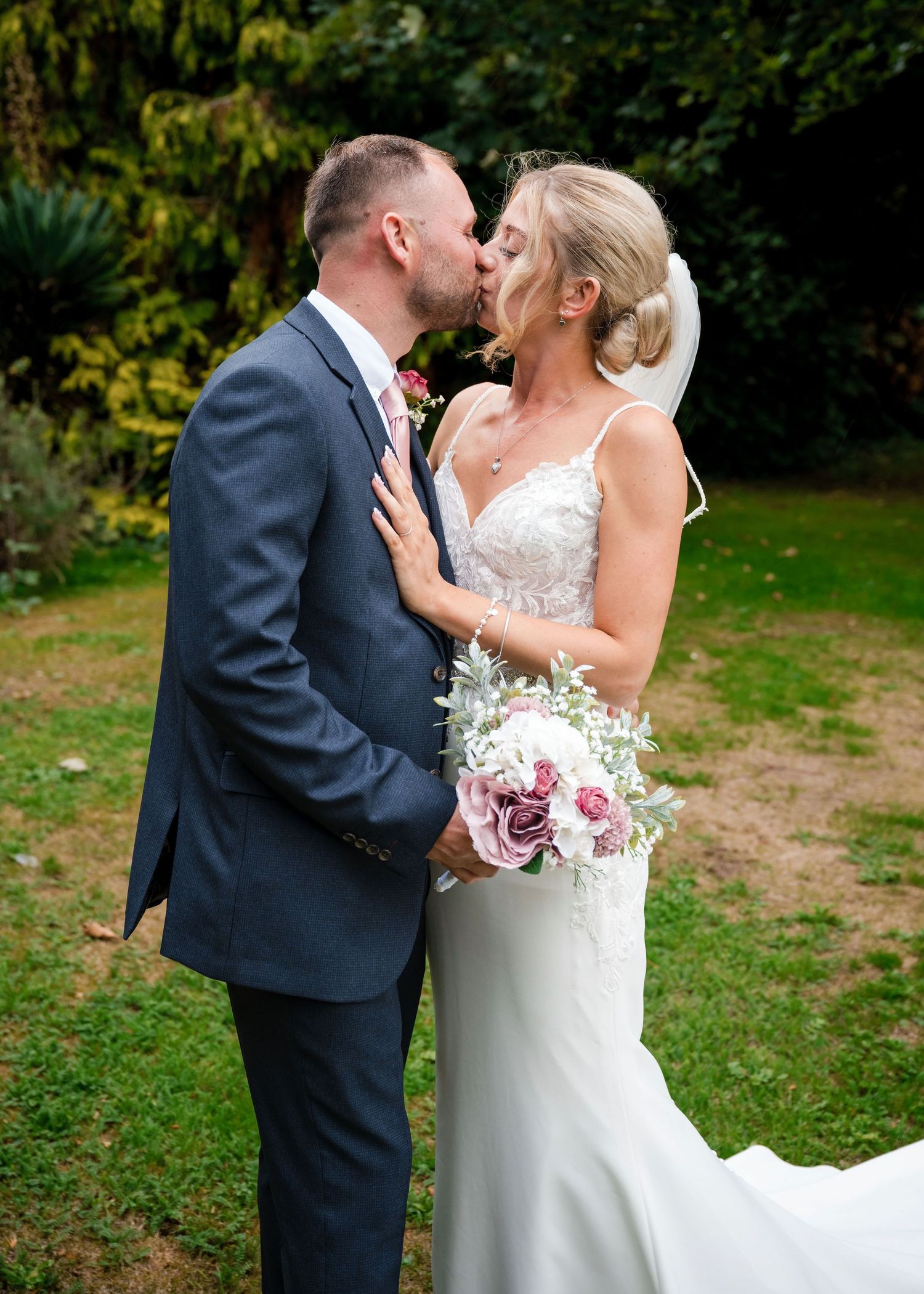 Bride and groom sharing a romantic kiss outdoors, bride holding a bouquet of pink roses.