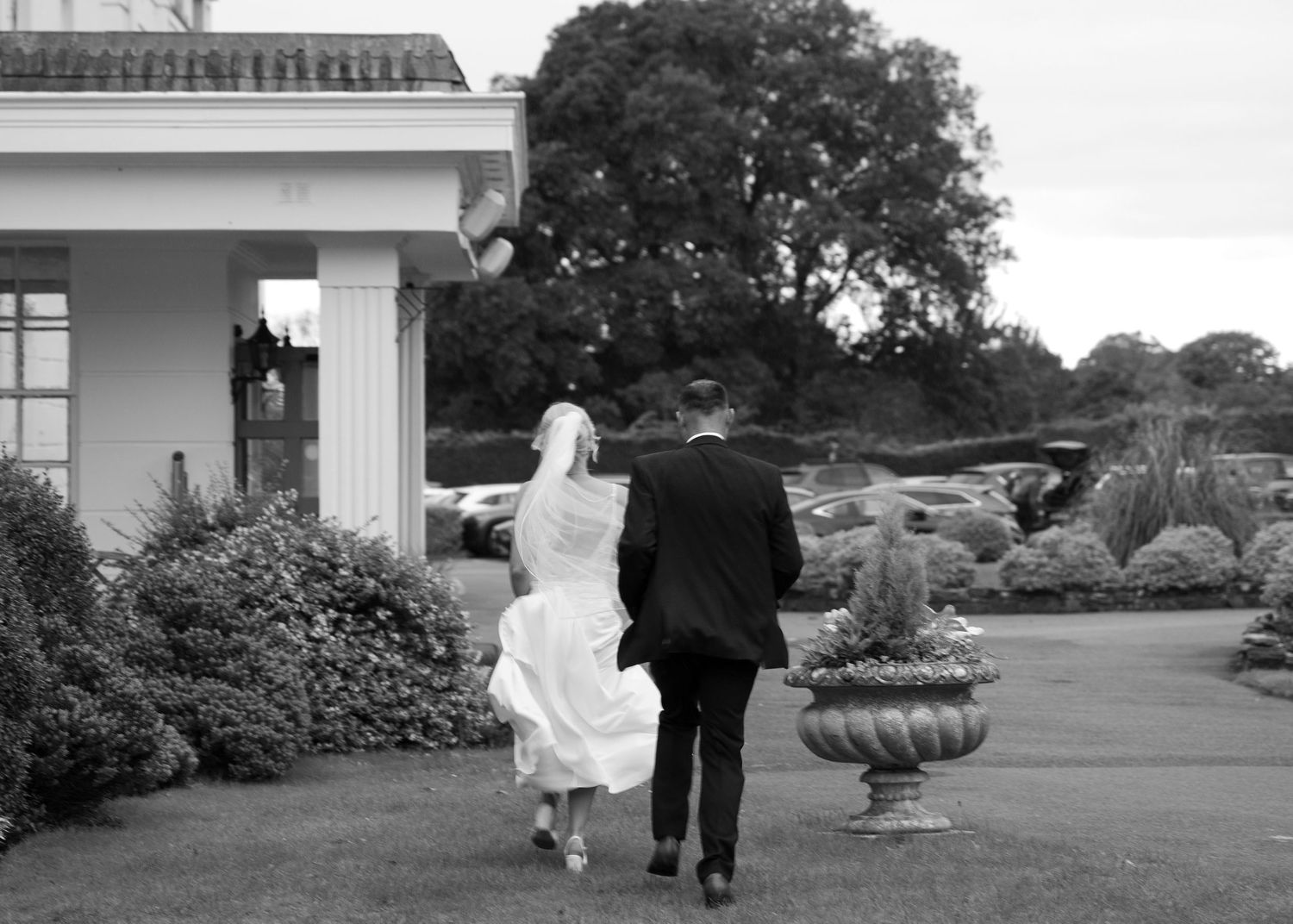 Black and white photo of a bride and groom walking hand in hand across a lush garden toward an elegant estate.