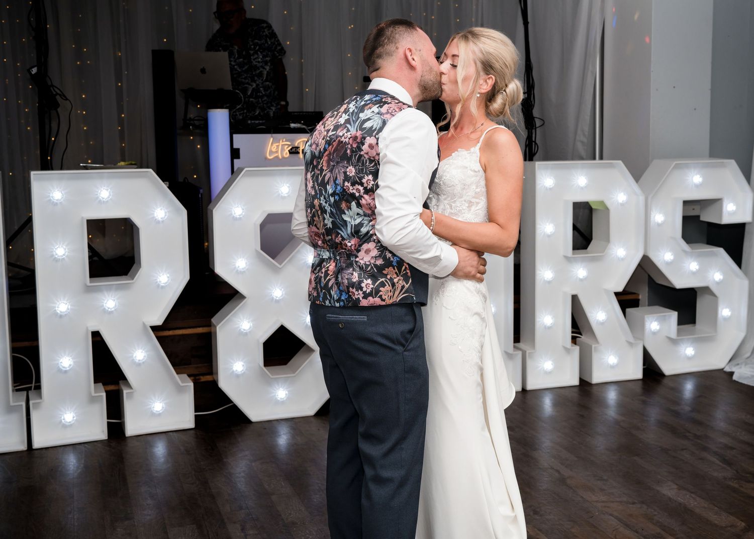 Newlyweds share a romantic first dance kiss in front of illuminated marquee letters at their wedding reception.