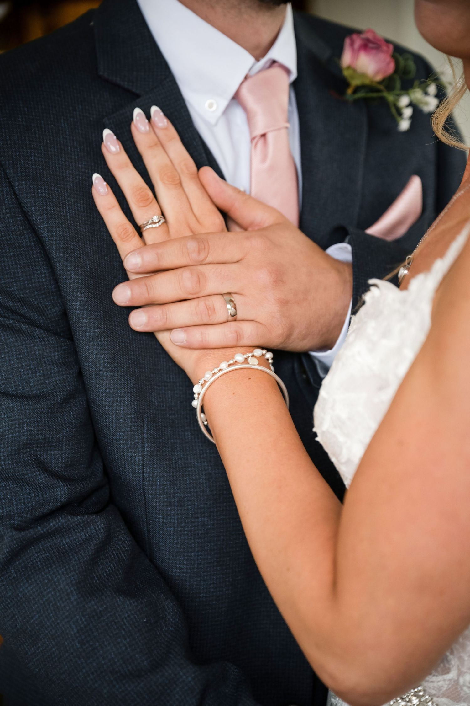 Bride and groom displaying wedding rings, bride wearing pearl bracelet and white lace dress, groom in gray suit with pink tie.