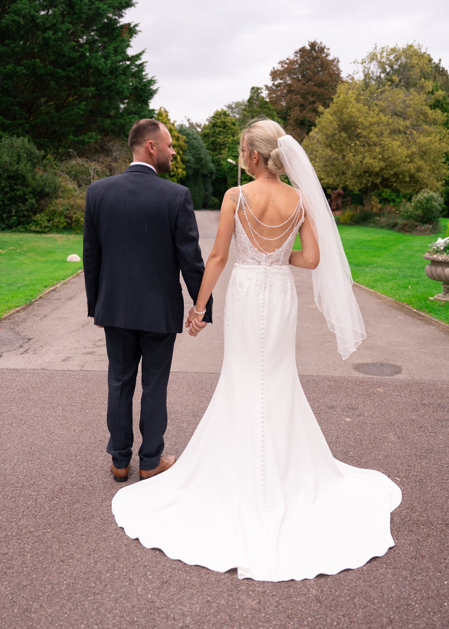 Bride and groom walking hand in hand down a path, bride wearing a backless white gown with veil and train.