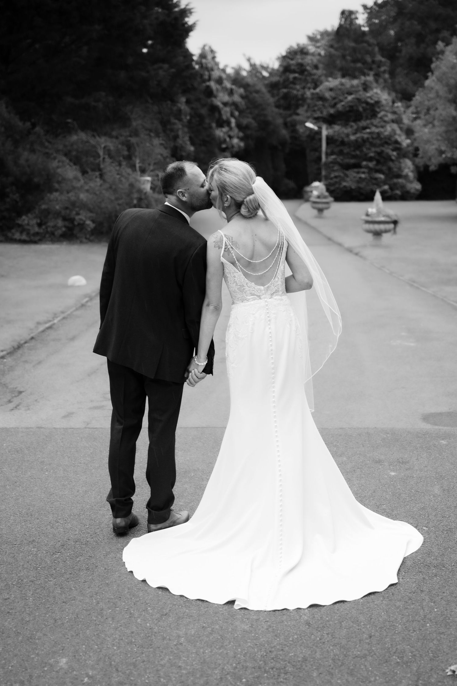 Black and white photo of bride and groom walking hand in hand, sharing a kiss, with a flowing wedding dress train.