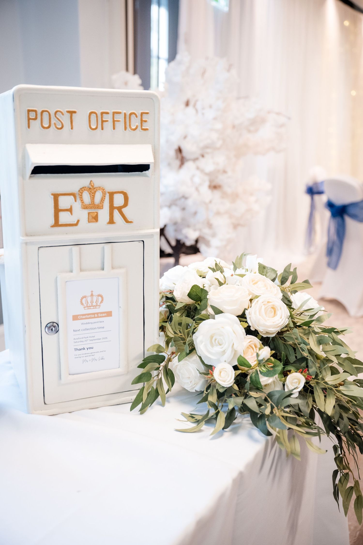 Elegant white Royal Mail post office wedding card box decorated with ER emblem beside white rose floral arrangement.