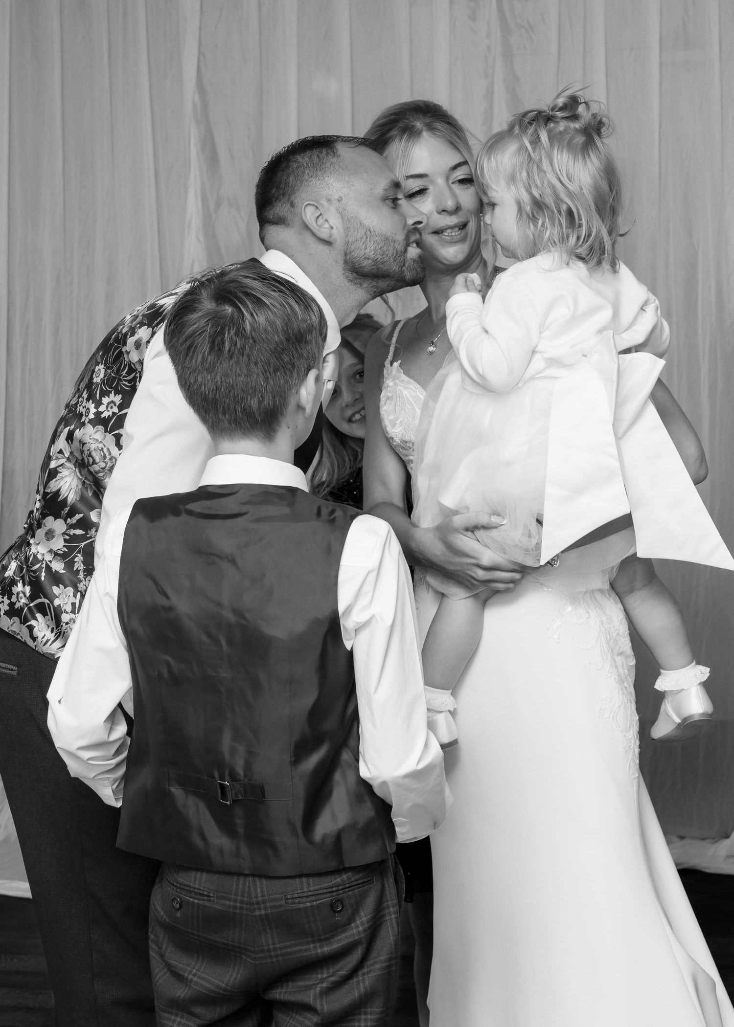 Groom kissing bride on cheek while she holds a young child, surrounded by wedding guests in black and white.