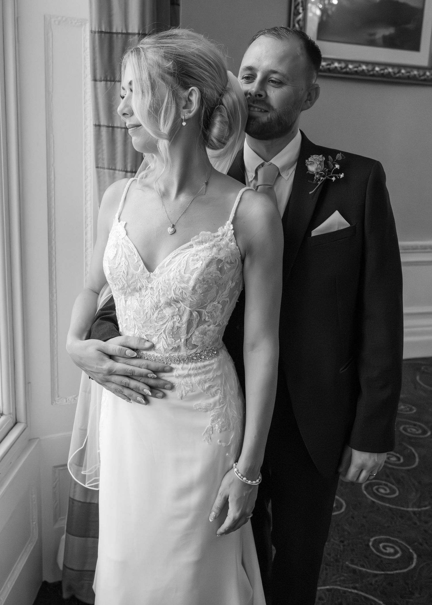 Black and white wedding portrait of bride in lace gown and groom in suit embracing by a window.