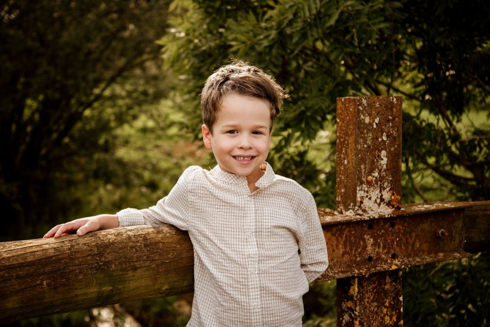 Smiling young boy leaning on a rustic wooden fence surrounded by lush green trees outdoors.