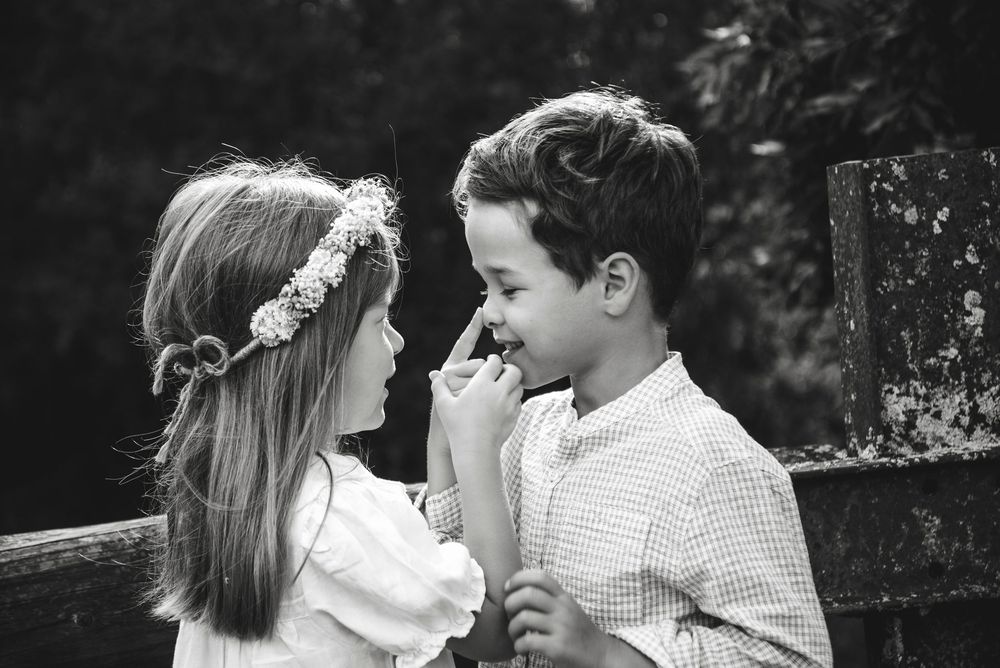 Black and white photo of two young children sharing a sweet moment, girl wearing floral crown, boy smiling warmly.