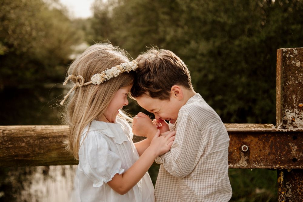 Two young children share a tender moment on a wooden bridge surrounded by lush greenery.