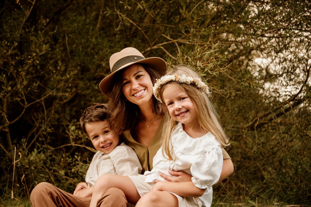 A smiling woman in a tan hat sits outdoors with two young children in white outfits, surrounded by lush greenery.