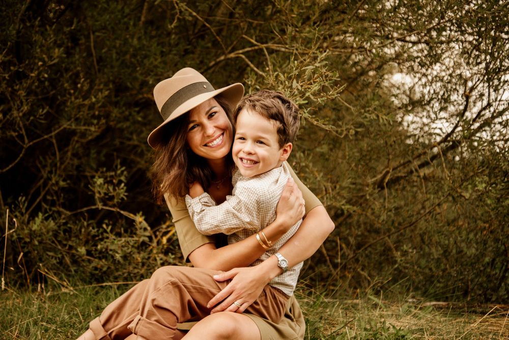 A smiling woman in a tan hat hugs a laughing boy outdoors among green trees in warm natural light.