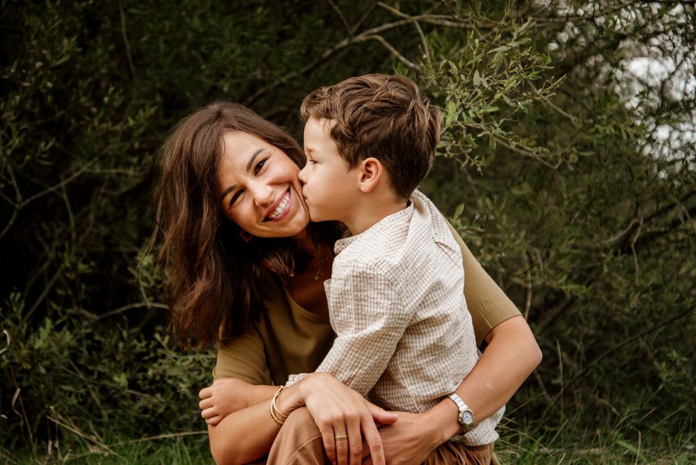A young boy lovingly kisses his smiling mother on the cheek outdoors surrounded by lush green foliage.