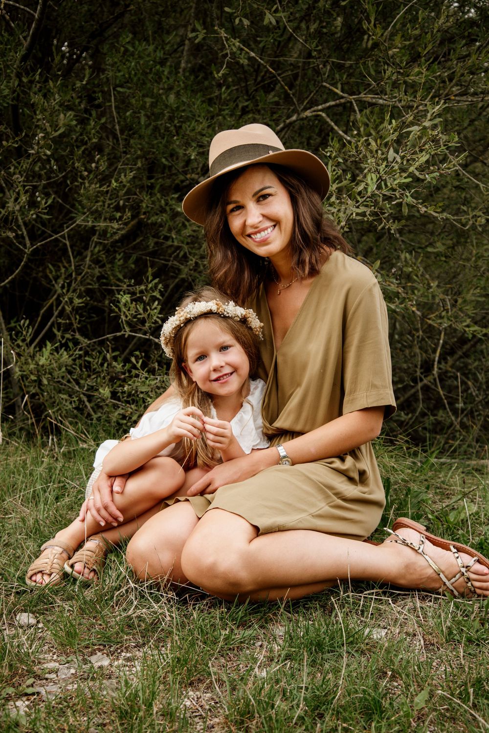 A smiling woman in a tan hat and olive dress sits outdoors with a young girl wearing a floral crown.